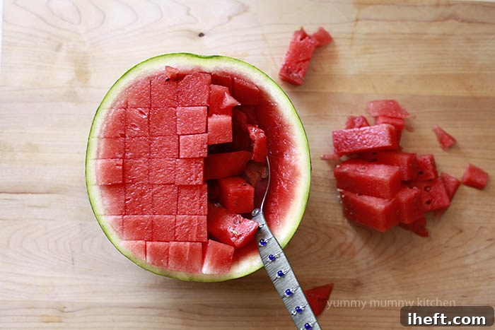Scooping out perfectly cut watermelon cubes from half of a watermelon with a spoon to create a natural serving bowl