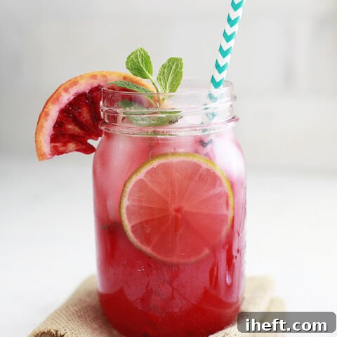 A red blood orange margarita in a mason jar garnished with lime and blood orange slices and a sprig of mint. The glass sits on a white kitchen counter.