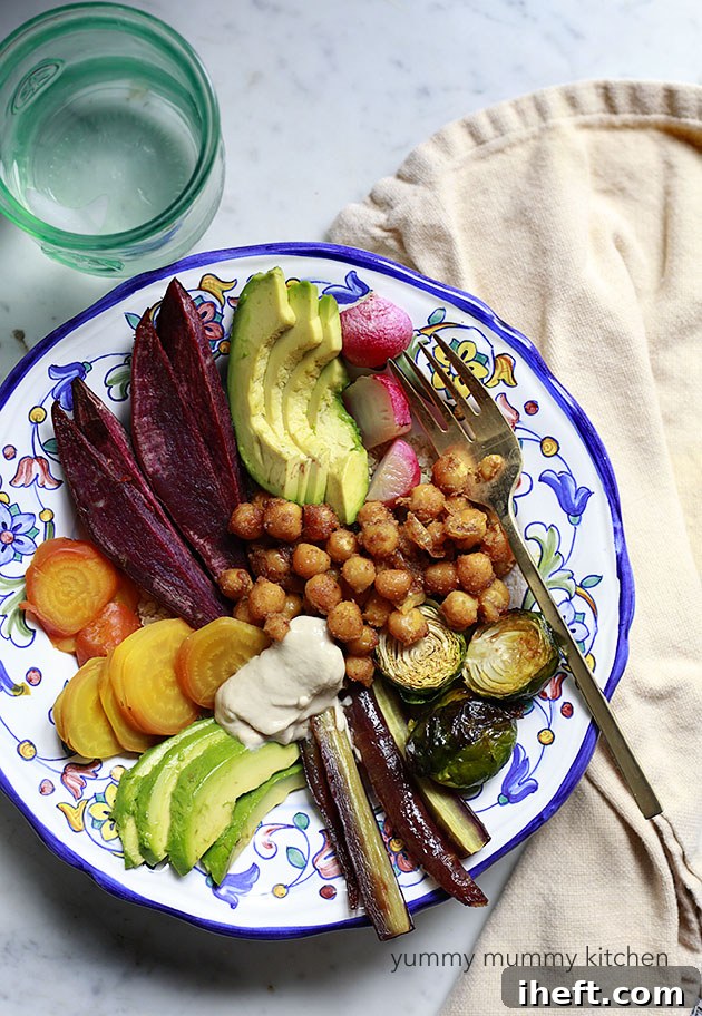 A vibrant vegan nourish bowl featuring perfectly roasted curried chickpeas, nestled among a medley of colorful root vegetables like Brussels sprouts, purple carrots, purple sweet potatoes, and golden beets, topped with creamy avocado and a drizzle of tahini over a bed of fluffy quinoa.
