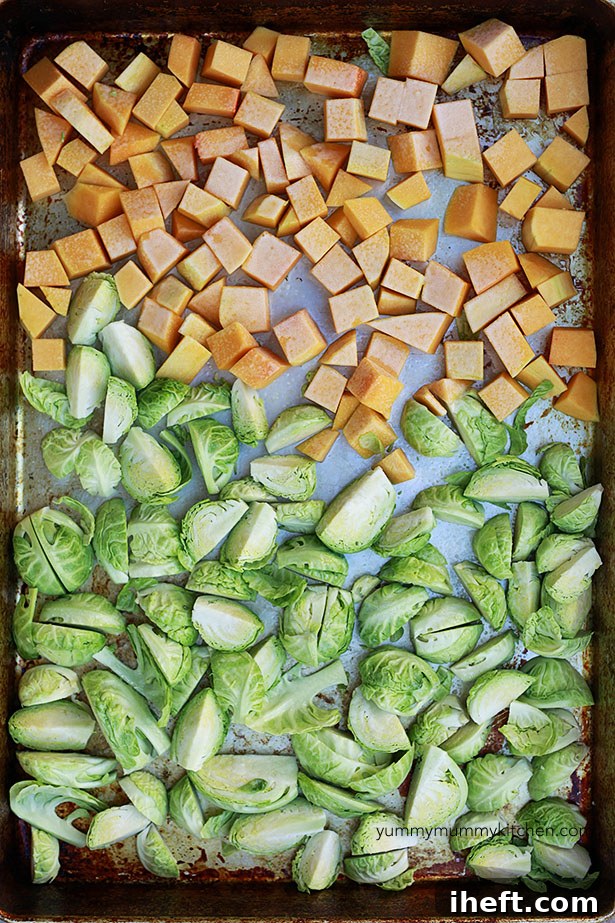 A large sheet pan filled with perfectly cubed butternut squash and quartered Brussels sprouts, seasoned and ready for roasting.