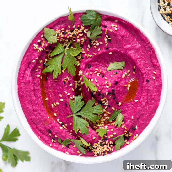 A close-up photo of pink beet hummus dip in a white bowl garnished with parsley and sesame seeds.