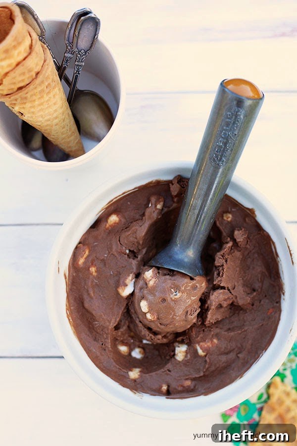 A beautifully presented bowl of creamy vegan rocky road ice cream, generously topped with mini vegan marshmallows and crunchy roasted almonds, with an ice cream scooper resting beside it. This easy dessert is made effortlessly in a blender, highlighting its homemade charm.