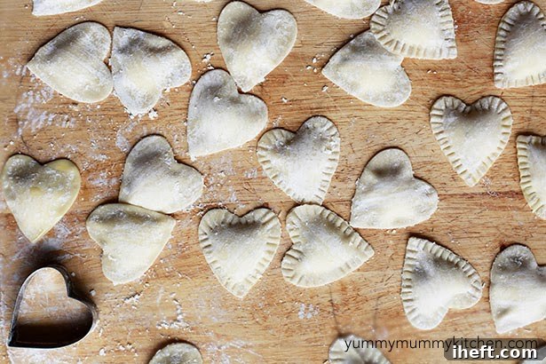 Heart shaped ravioli made with wonton wrappers on a cutting board with a heart-shaped cookie cutter on the side. 