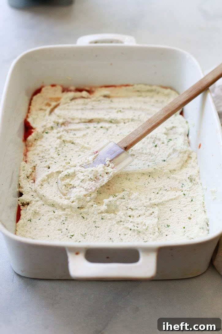 A hand smoothly spreading a generous layer of creamy vegan almond ricotta, flecked with fresh parsley, over lasagna noodles and vegetables in a baking dish, showcasing a delicious stage in the preparation of this dairy-free lasagna.