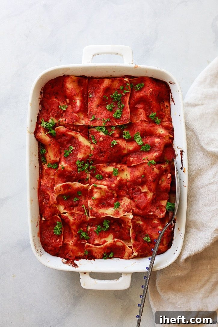 A baked white casserole dish of vegan lasagna, neatly cut into squares and garnished with fresh parsley, ready to be served. The top layer is golden and bubbly, hinting at the warm, comforting flavors within.