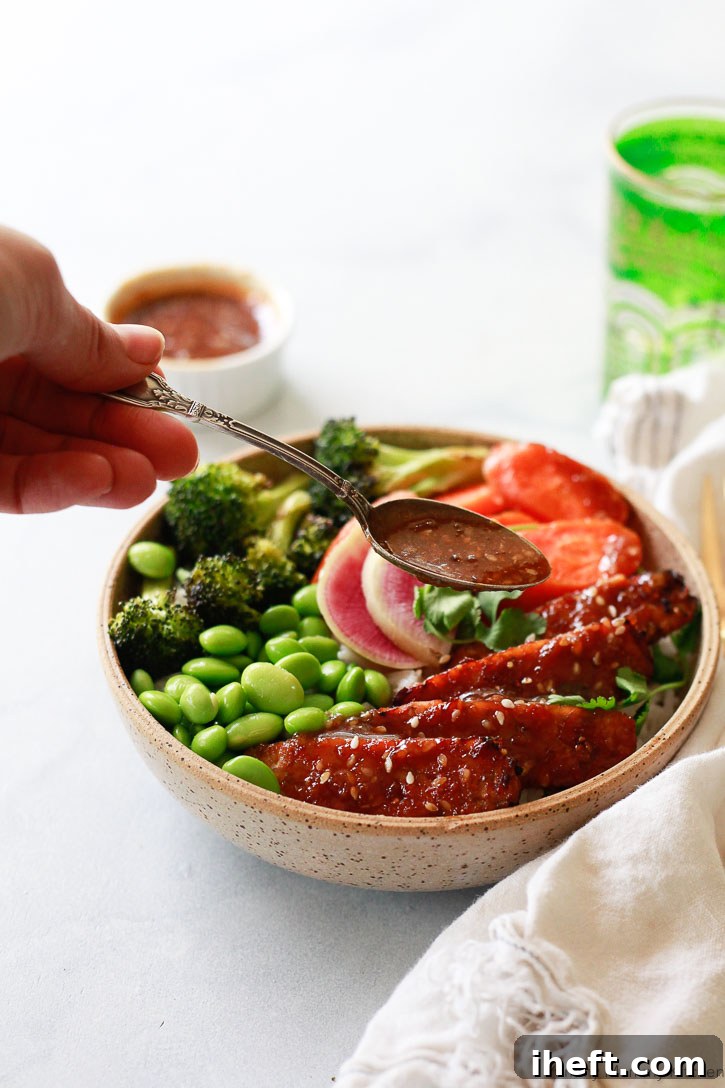 A beautifully arranged bowl featuring homemade sesame seed teriyaki sauce drizzled generously over teriyaki-glazed tempeh, fluffy rice, vibrant edamame, crisp broccoli, and tender carrots, making a wholesome and appealing meal.