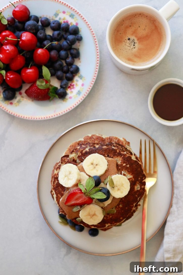 A beautifully arranged vegan pancake breakfast featuring a stack of banana pancakes topped with fresh blueberries and almond butter, accompanied by a side of vibrant cherries, more blueberries, a small cup of maple syrup, and a steaming mug of coffee, ideal for a special morning like Mother's Day.