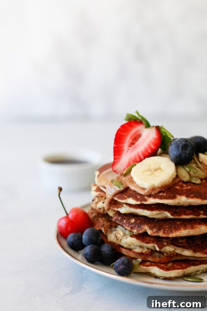 A tantalizing close-up shot of easy one-bowl vegan banana pancakes made with oats and chia seeds, garnished with fresh blueberries, strawberries, and pepitas, featuring a luscious drizzle of almond butter cascading down the sides, emphasizing their deliciousness and plant-based appeal.