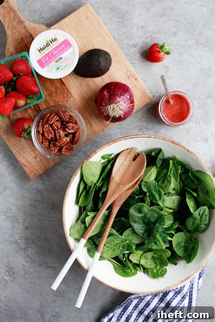 All the essential ingredients for a delicious strawberry spinach salad are neatly arranged on a sleek grey countertop. This inviting display includes a bowl of fresh baby spinach, vibrant whole strawberries, creamy avocado, crunchy pecans, soft cheese, and a jar of homemade strawberry dressing, ready for assembly.