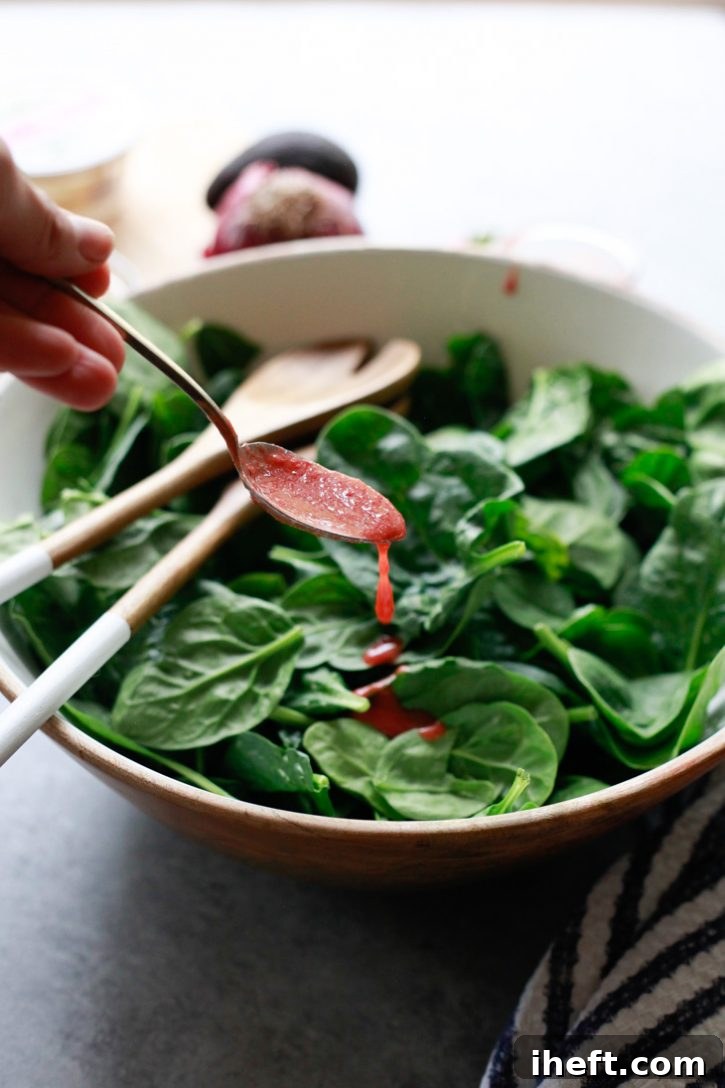 A bowl of fresh baby spinach is being gently tossed and dressed with a generous amount of homemade strawberry vinaigrette salad dressing, ensuring every leaf is coated.