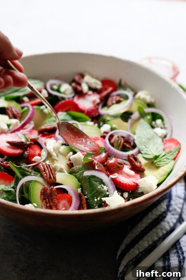 A luscious strawberry spinach salad, adorned with crisp red onions, creamy avocado, crumbled feta, and crunchy pecans, receives a final elegant drizzle of the homemade strawberry vinaigrette, enhancing its appeal and flavor.