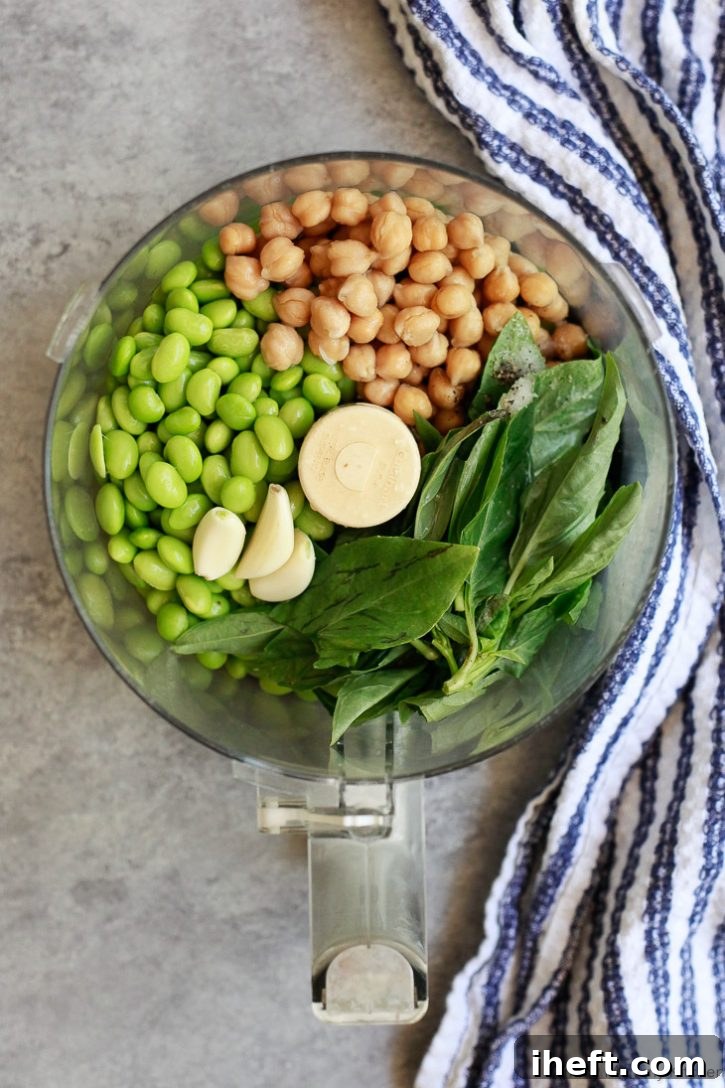 Chickpeas, edamame, basil, and garlic in the bowl of a food processor ready to make homemade edamame hummus. Fresh ingredients for a vibrant dip.