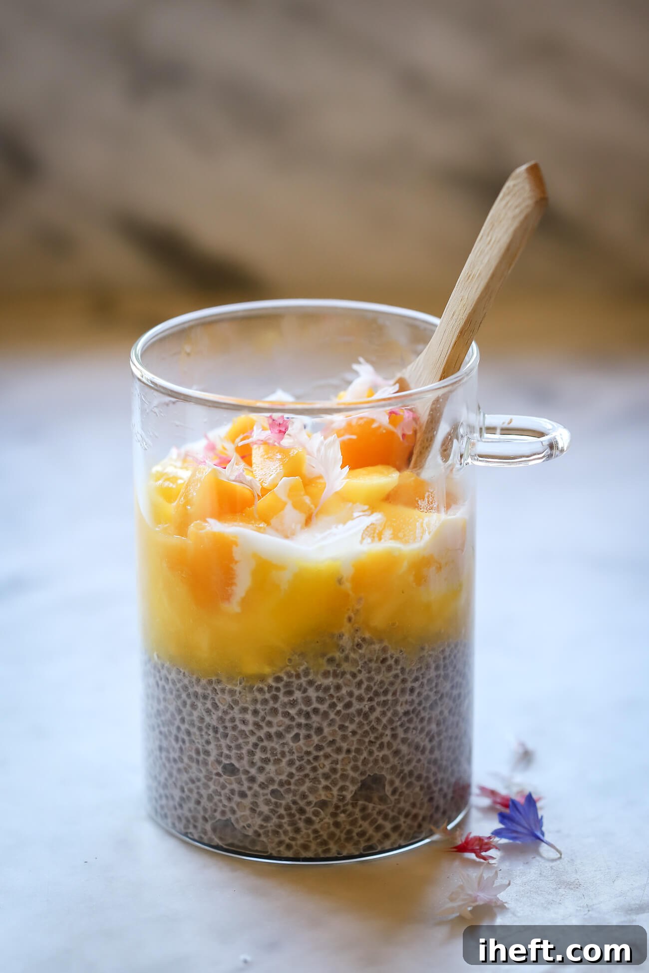 A beautifully layered glass jar of mango chia pudding, topped with fresh mango chunks, creamy yogurt, and delicate edible flowers, with a bamboo spoon attached, set on a bright marble surface.