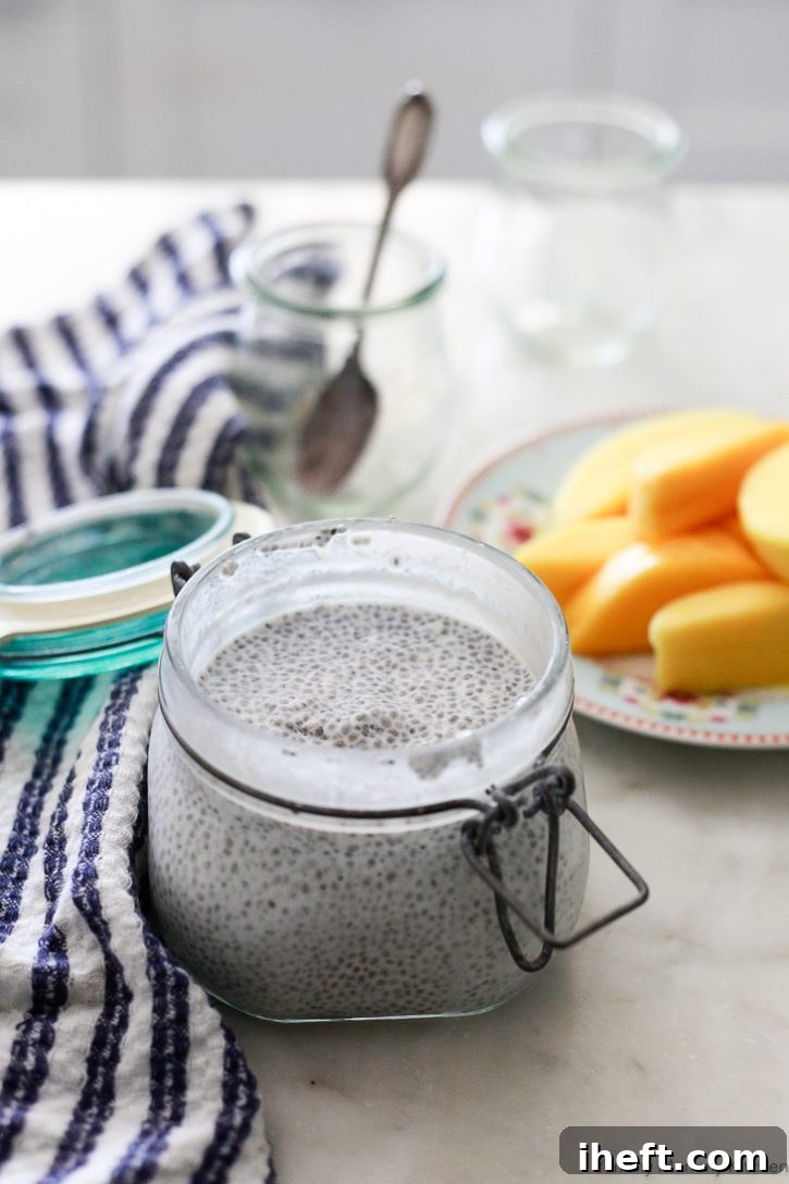 A tall clear glass jar is filled with creamy, pale yellow coconut chia seed pudding. In the background, a ripe sliced mango is arranged on a plate next to two smaller empty serving jars, hinting at the next steps of assembly.