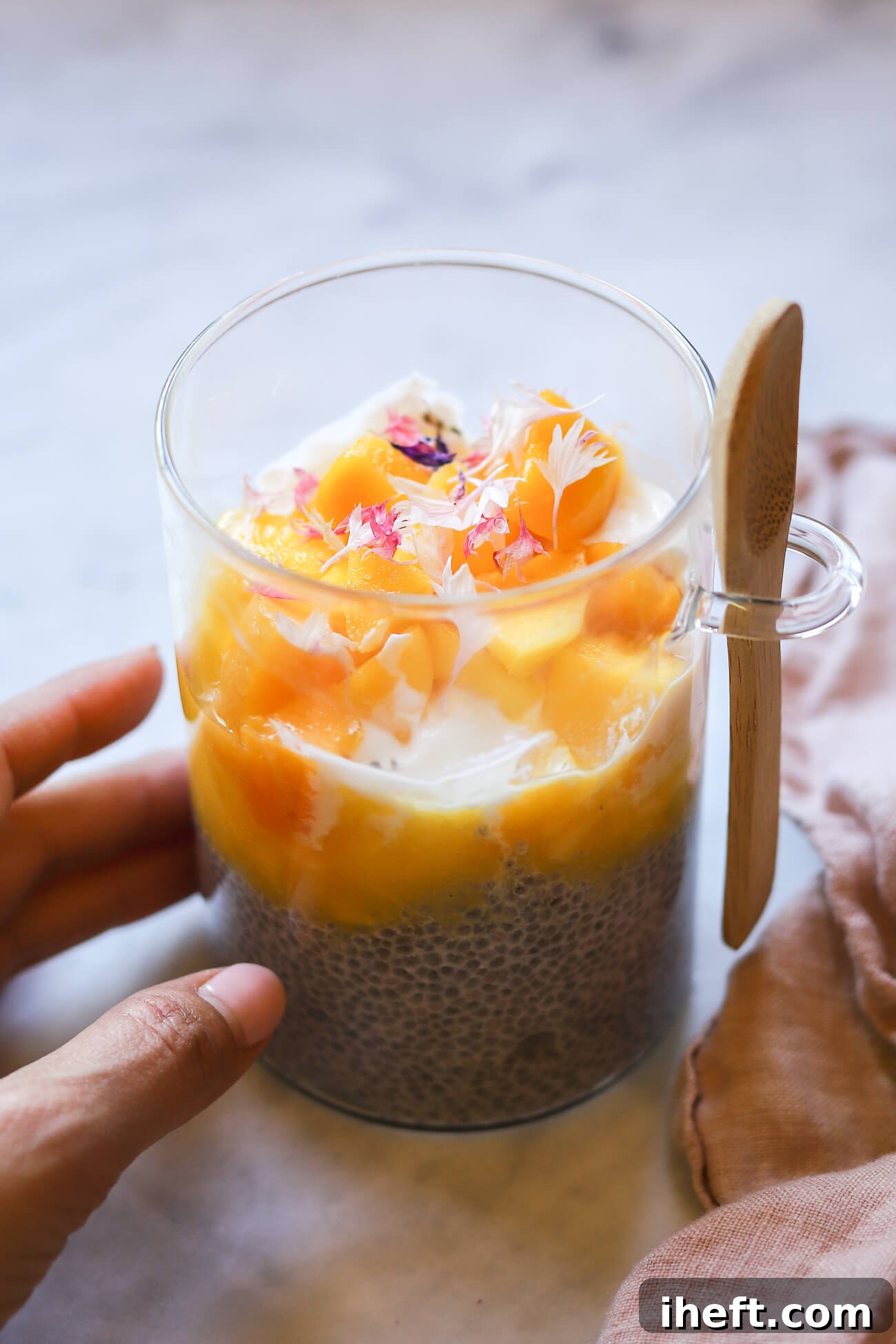 A hand reaches in to pick up a clear glass meal-prep jar, beautifully layered with mango chia pudding, creamy yogurt, and vibrant mango puree and chunks, all crowned with delicate edible pink bachelor button flowers. It's a perfect grab-and-go healthy meal.