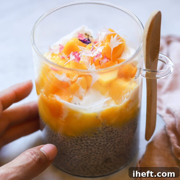 A glass filled with chia pudding topped with yogurt and mango puree and mango chunks with pink edible flowers. The meal-prep glass has a wooden spoon attached.