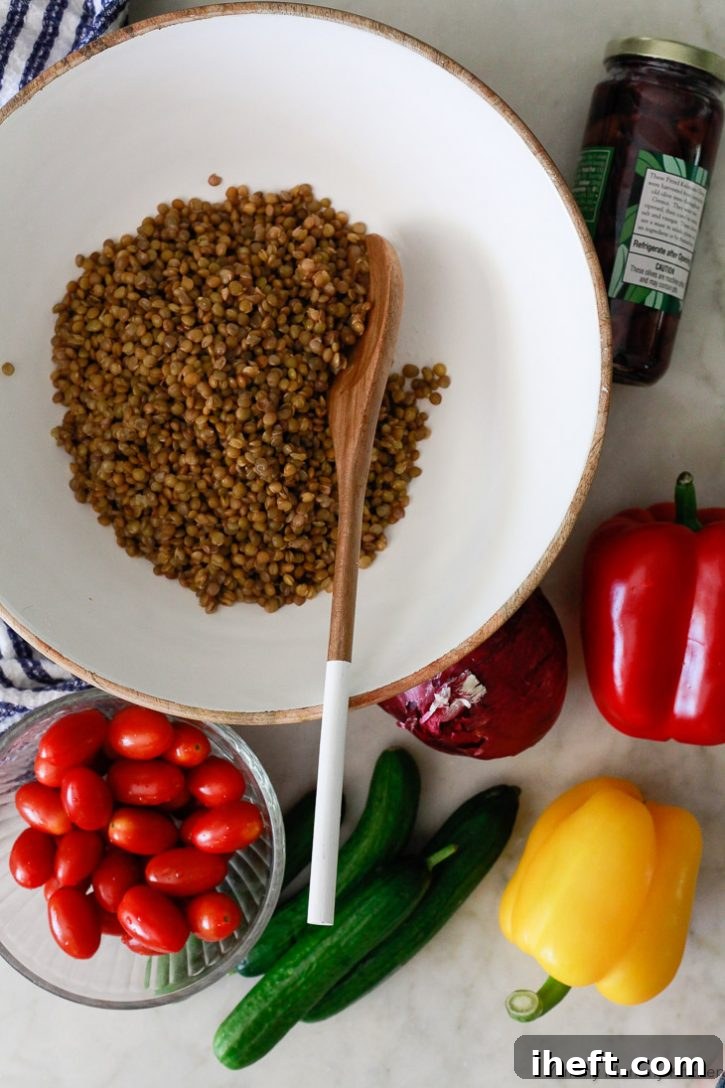 Mediterranean lentil salad ingredients on a white countertop include cooked green lentils, tomatoes, cucumber, bell peppers, and olives. 