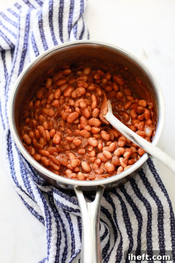 A beautiful saucepan of vegetarian baked beans rests on a tea towel, accompanied by a wooden spoon. This easy, vegan, and gluten-free recipe promises deliciously rich and flavorful beans.