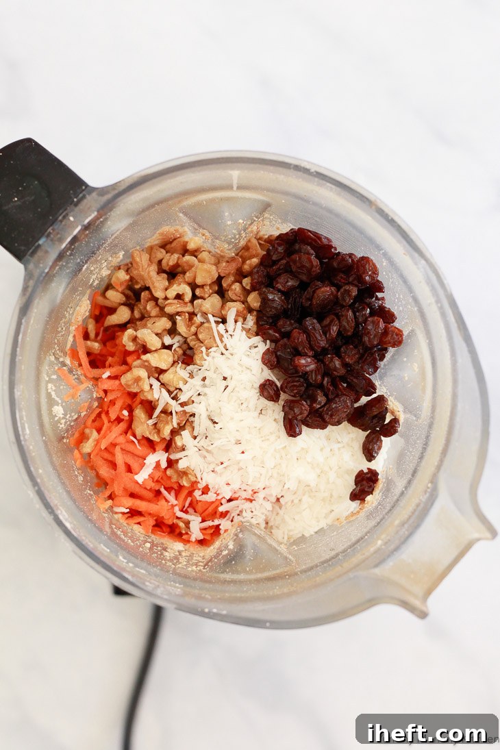 Gluten-free carrot muffins batter being prepared in a blender. Shredded carrots, chopped walnuts, sweet raisins, and shredded coconut are ready to be folded in.
