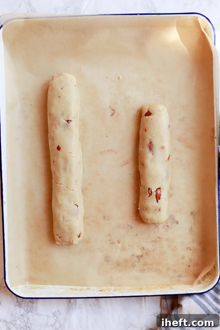 Two logs of gluten-free almond biscotti dough rest on a parchment-lined cookie sheet, prepared for their first bake.