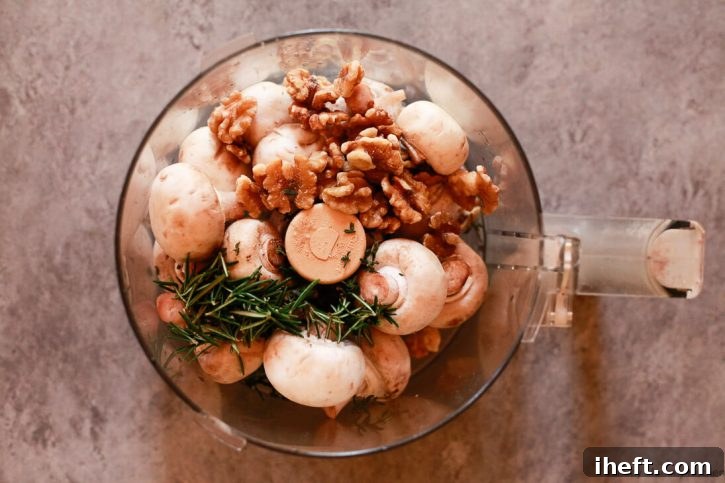 A food processor bowl filled with button mushrooms, walnuts, fresh thyme, and rosemary, ready to be pulsed into a fine, flavorful duxelles mixture for the vegetarian Wellington.