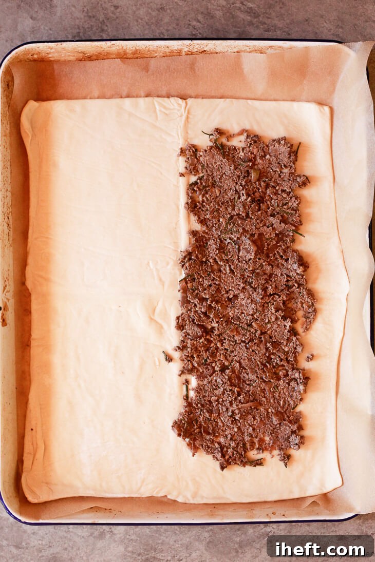 A sheet of thawed puff pastry expertly spread with a layer of mushroom duxelles, leaving a neat border for sealing, demonstrating the initial step in assembling a delicious vegetarian Wellington.