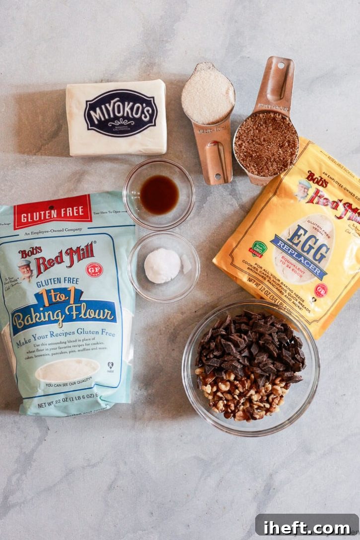 The ingredients for vegan chocolate chip cookies laid out on a marble countertop: gluten free baking flour, Miyoko's vegan butter, Bob's Red Mill Egg Replacer, brown and white sugar, vanilla extract, baking soda, vegan chocolate chunks, and walnuts.