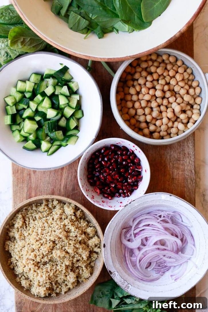 A display of fresh ingredients for chickpea quinoa salad on a rustic cutting board: cooked quinoa, vibrant spinach, plump chickpeas, chopped cucumber, glistening pomegranate arils, and thinly sliced red onion.