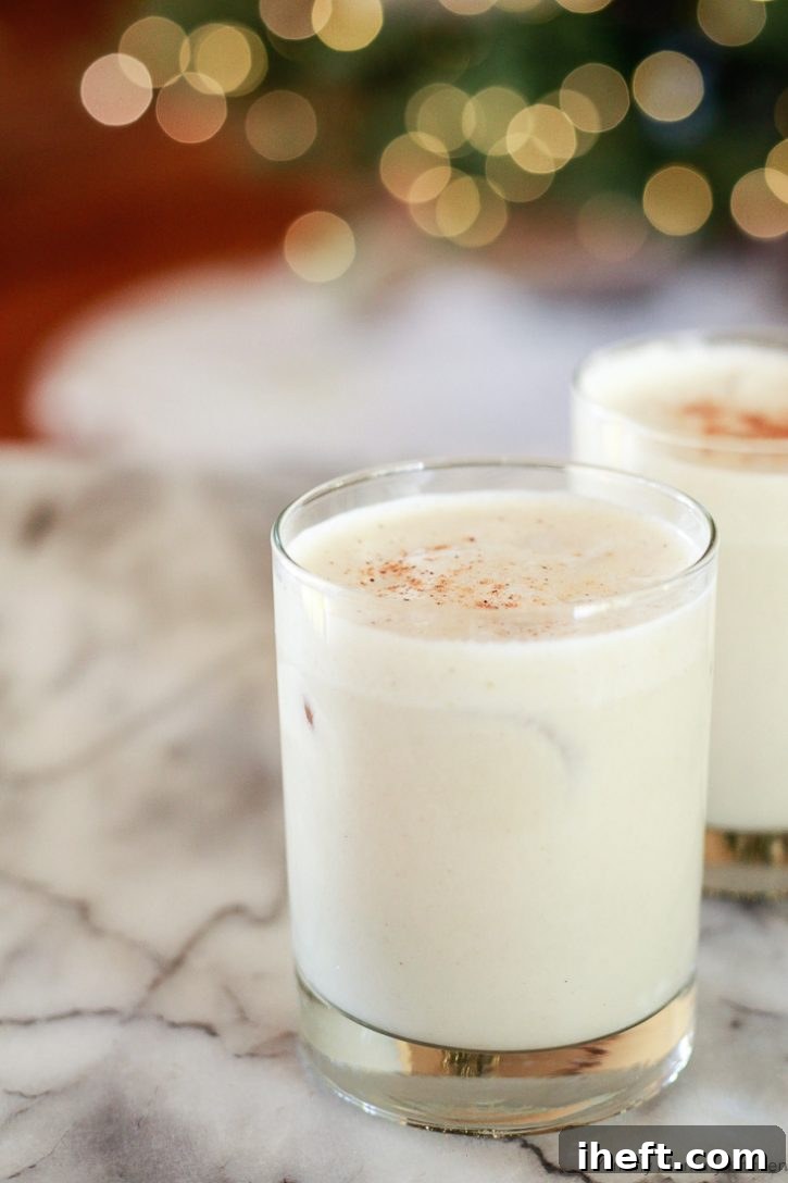 Glasses of vegan eggnog in front of bokeh Christmas tree lights. This creamy cold vegan nutmeg is made with almond and coconut milk, sweetened with dates, and topped with nutmeg.