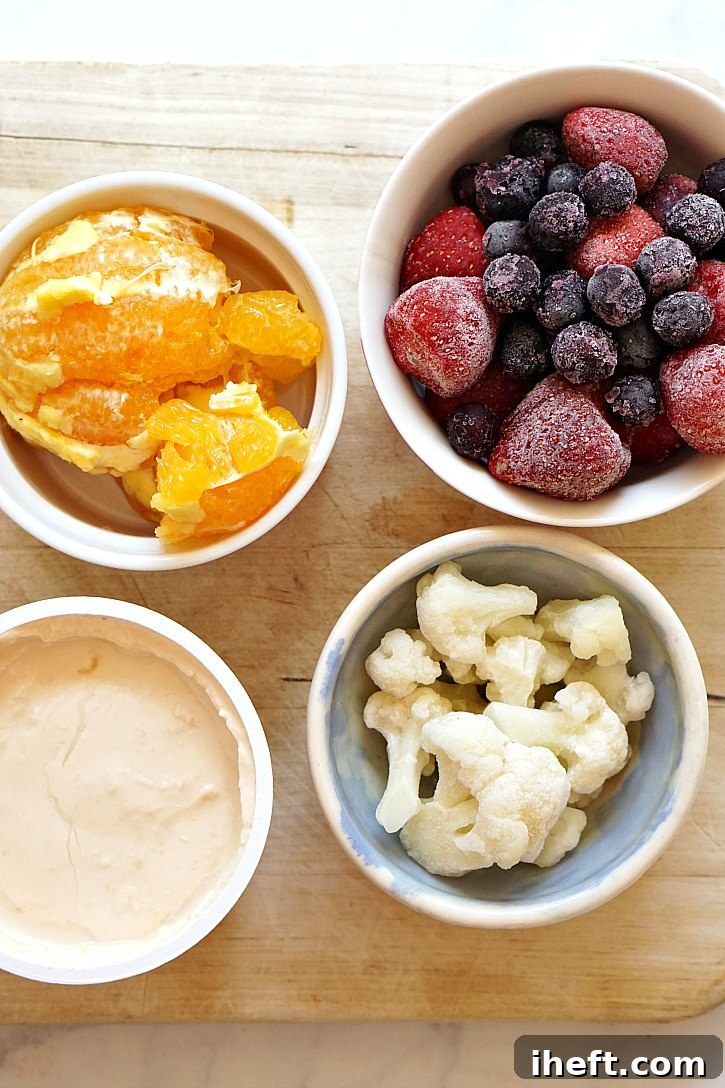 All the fresh and frozen ingredients laid out beautifully on a wooden surface, ready for blending in a high-speed Vitamix. This healthy vegan cauliflower smoothie includes vibrant blueberries, sweet strawberries, a peeled orange, frozen cauliflower florets, and a creamy plant-based yogurt.