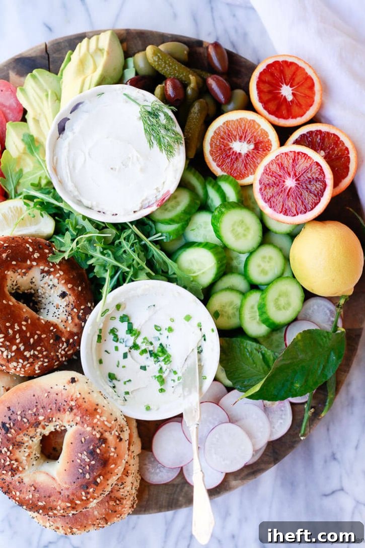A beautiful breakfast bagel platter with two bowls of homemade vegan cream cheese with chives, cucumber, blood oranges, radish, avocado and olives on a round cutting board. 