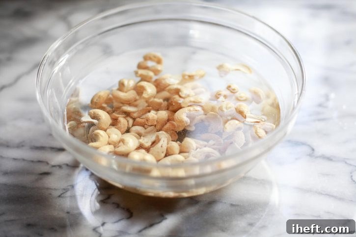 Raw cashews soak in water in a bowl on a marble countertop as the first step in making homemade vegan cream cheese. 
