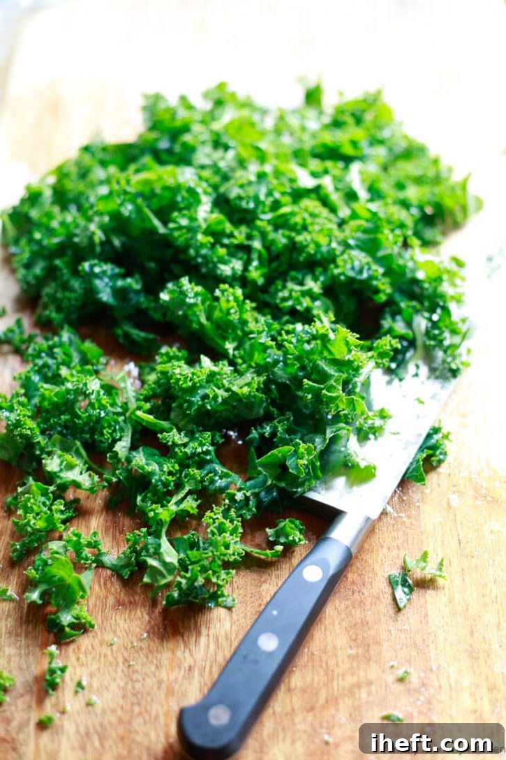 Fresh, bright green curly kale leaves, meticulously chopped into bite-sized pieces, await their addition to a simmering pot of white bean and kale soup on a rustic wooden cutting board.