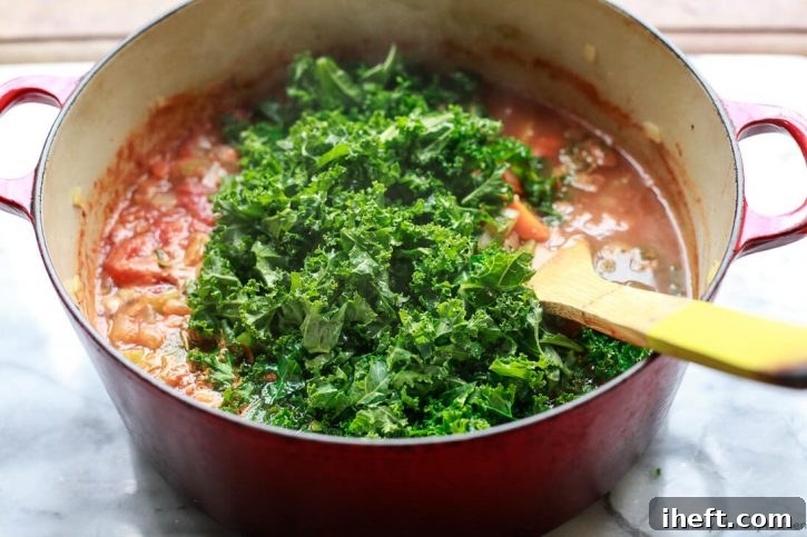 Vibrant chopped green kale being stirred gently into the rich, red broth of white bean and kale soup in a large red Dutch oven, adding freshness and nutrients.