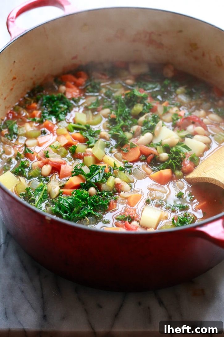 A close-up view of a large, red Dutch oven brimming with hot, steaming white bean and kale soup, showcasing its inviting texture and warmth.
