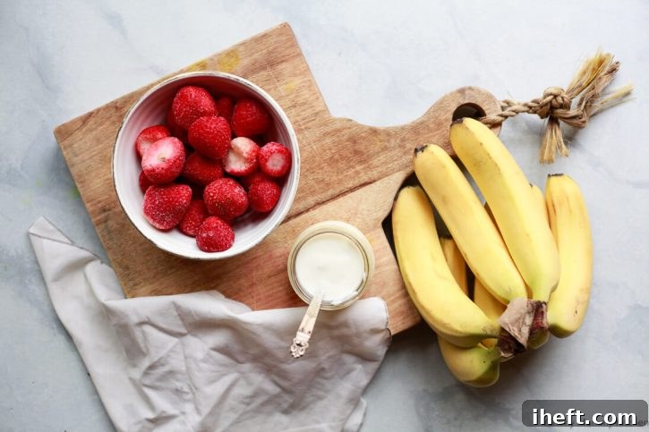 A bowl of frozen strawberries, a cup of yogurt, and a bunch of bananas sit on a white countertop with a cutting board. These are ingredients for a strawberry banana smoothie.