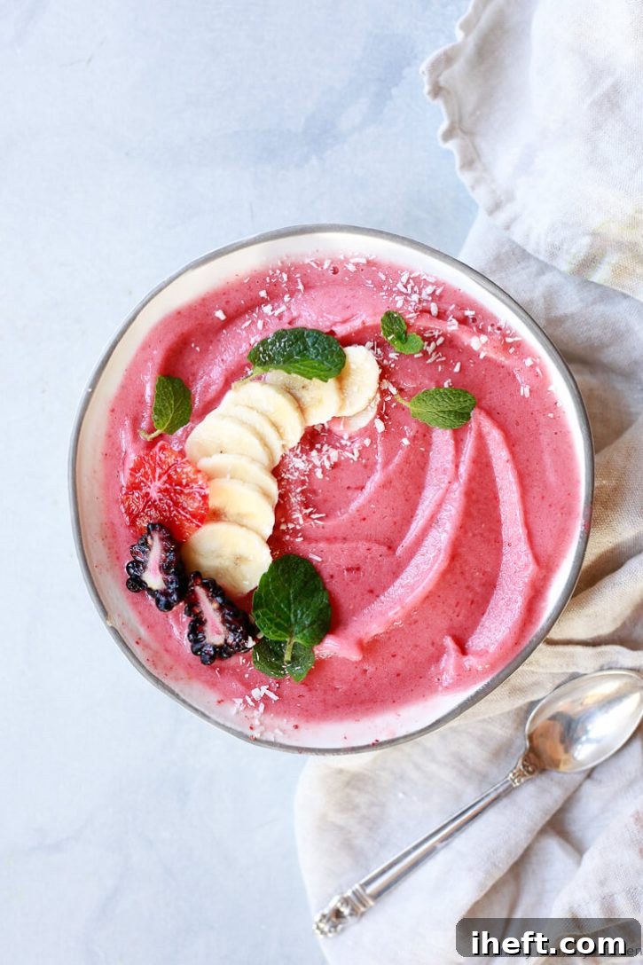 A beautiful smoothie bowl filled with a strawberry banana smoothie and topped with sliced banana, blackberries, orange, and mint leaves with a sprinkle of coconut shreds. 