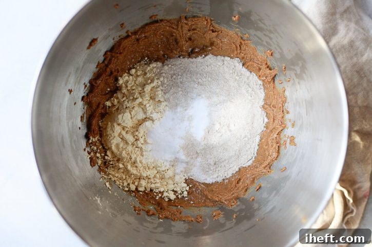 A mixing bowl filled with the whipped peanut butter base, now topped with carefully measured whole wheat flour, chickpea flour, and baking soda, ready to be combined for the peanut butter oatmeal snack squares.