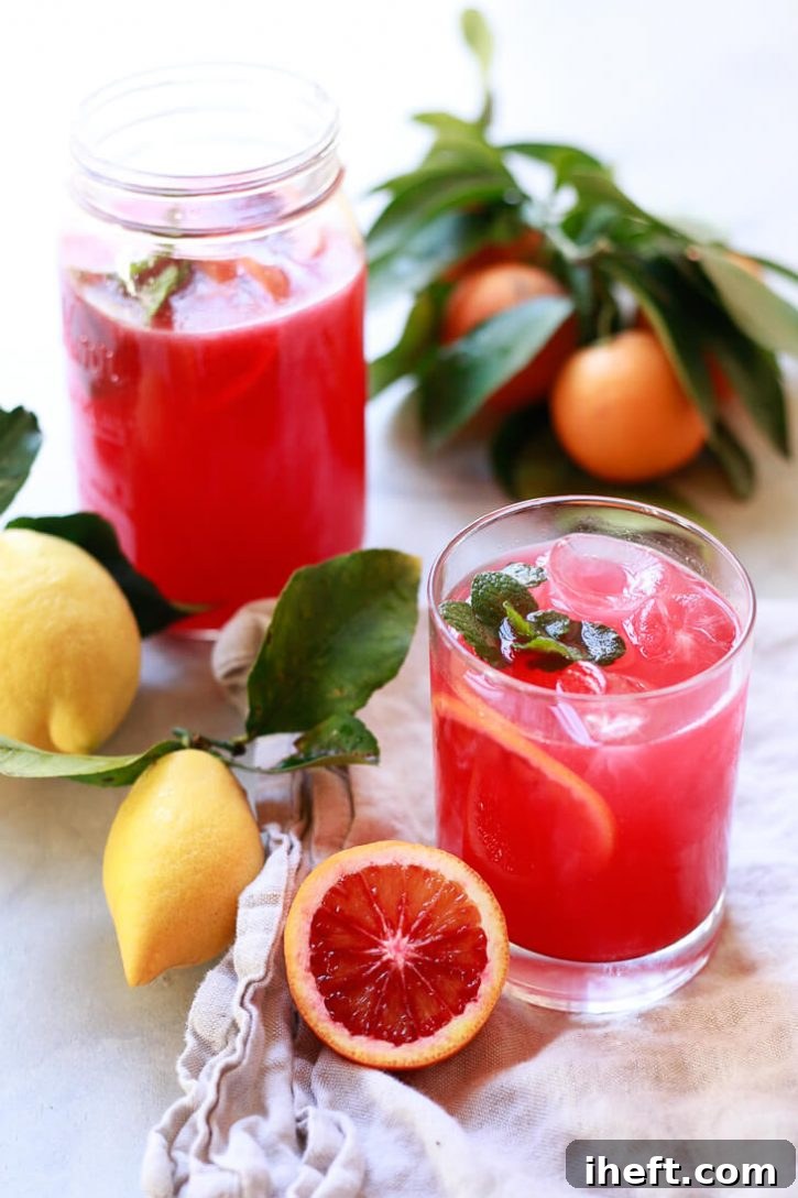 A glass of blood orange lemonade sits on a counter with blood oranges and lemons. 