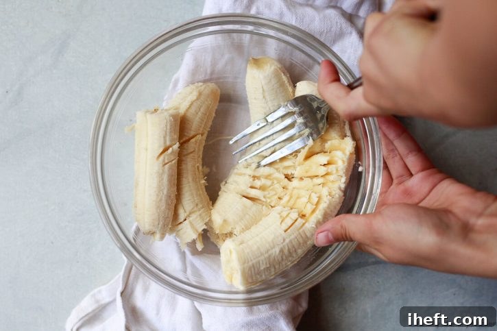 A child's hands mash bananas with a fork in a glass bowl.