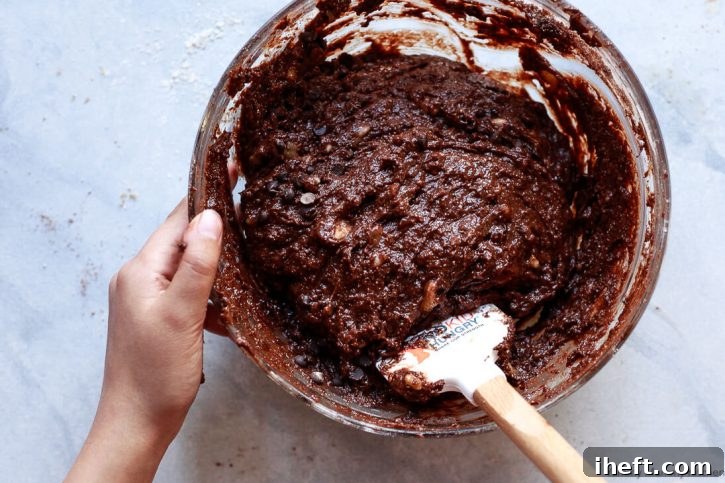 A child stirs together chocolate banana muffin batter in a glass bowl on a white countertop.