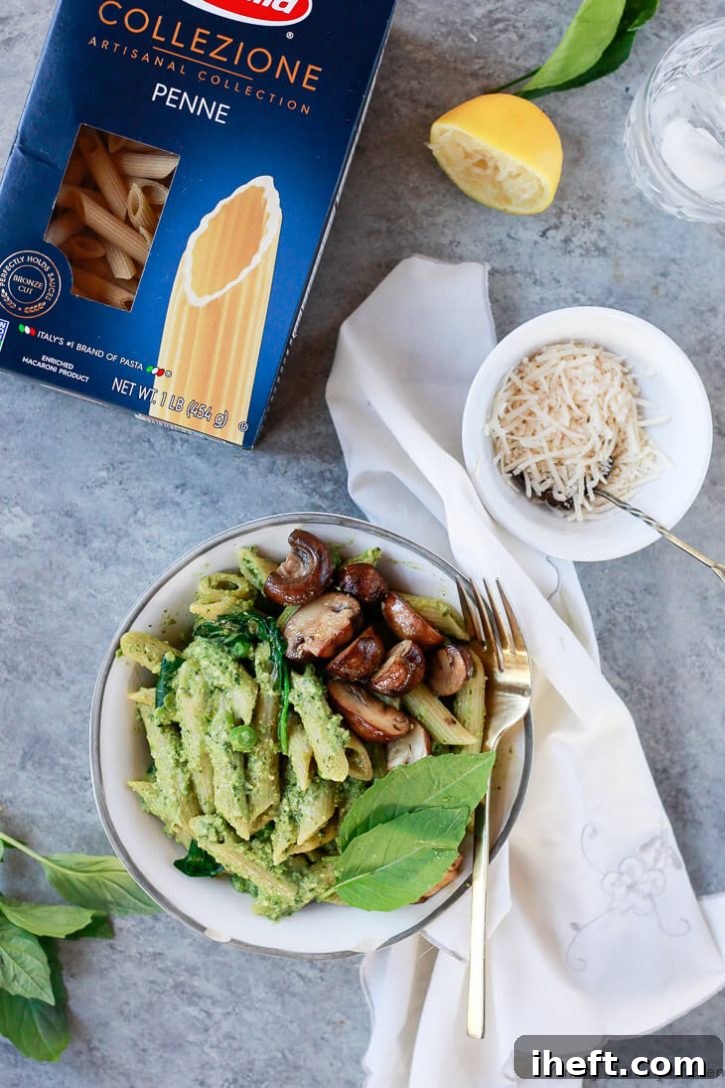 A bowl of vegan pea pesto pasta with mushrooms and spinach sit on a grey countertop with a bowl of vegan cheese. 