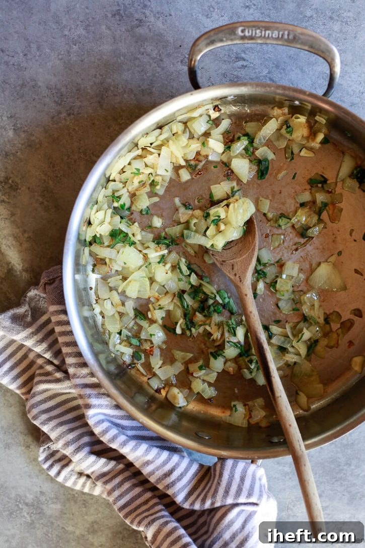 Finely diced onions, thinly sliced garlic, and fresh oregano sprigs gently sautéing in shimmering olive oil in a medium pan, stirred with a wooden spoon.