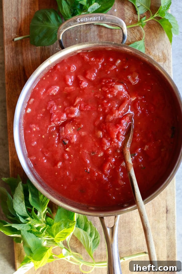 A pan filled with freshly combined pomodoro sauce ingredients, including crushed tomatoes, onions, garlic, and herbs, before it begins to simmer on the stove. Fresh basil leaves are artfully placed on a wooden cutting board nearby.
