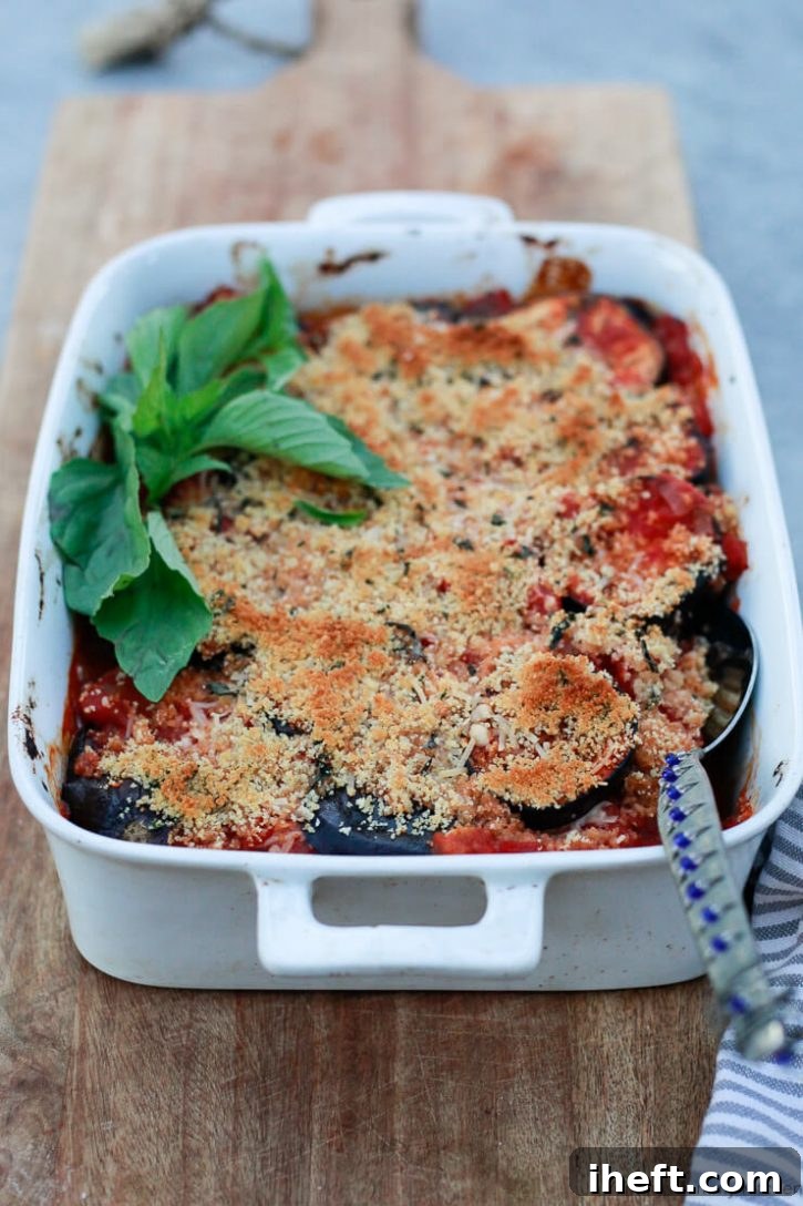 A white casserole dish filled with baked vegan eggplant parmesan, topped with golden brown breadcrumbs and garnished with fresh basil. 
