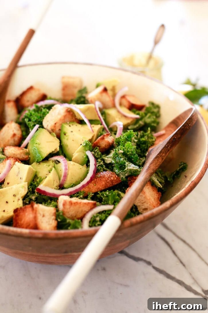 A large, rustic wooden bowl is overflowing with a vibrant vegan kale Caesar salad, generously mixed with golden croutons, creamy avocado slices, and delicate red onion slices. The salad is a colorful and appetizing medley of fresh ingredients, promising a delightful plant-based meal.