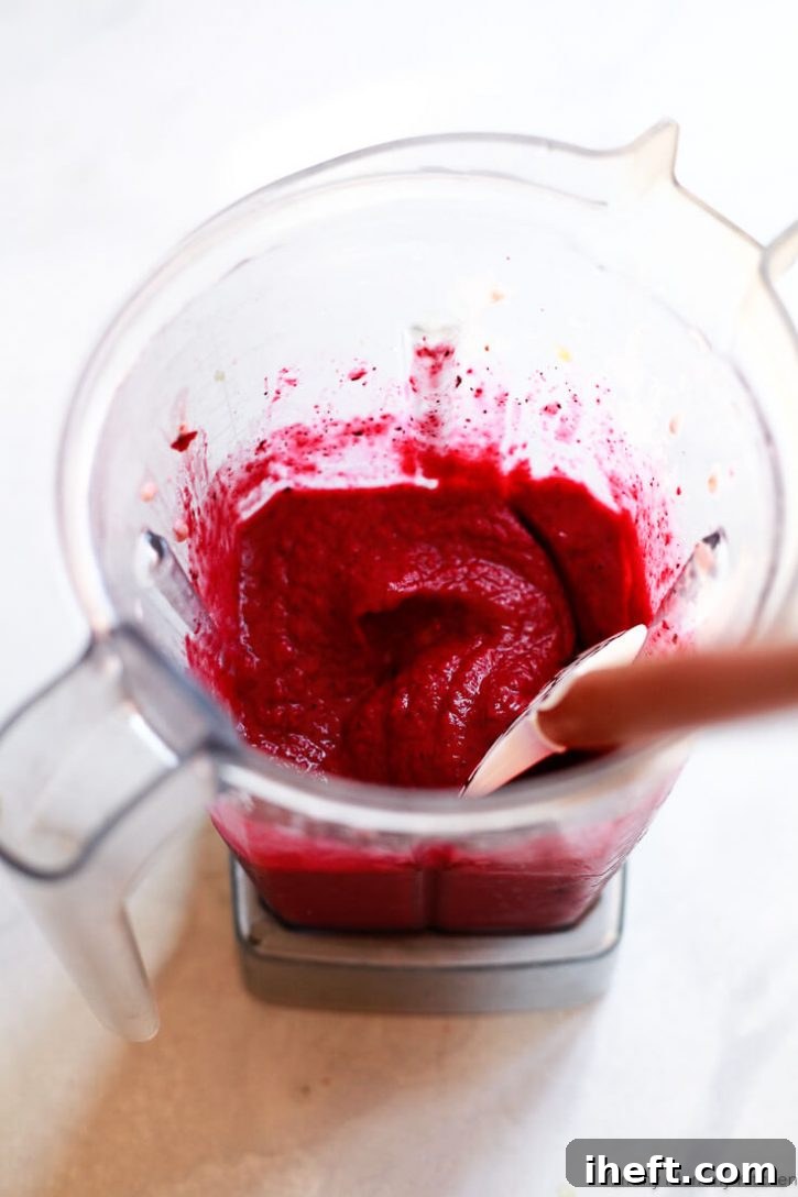 A bright red smoothie blended in a Vitamix blender gets scooped out with a rubber spatula. 