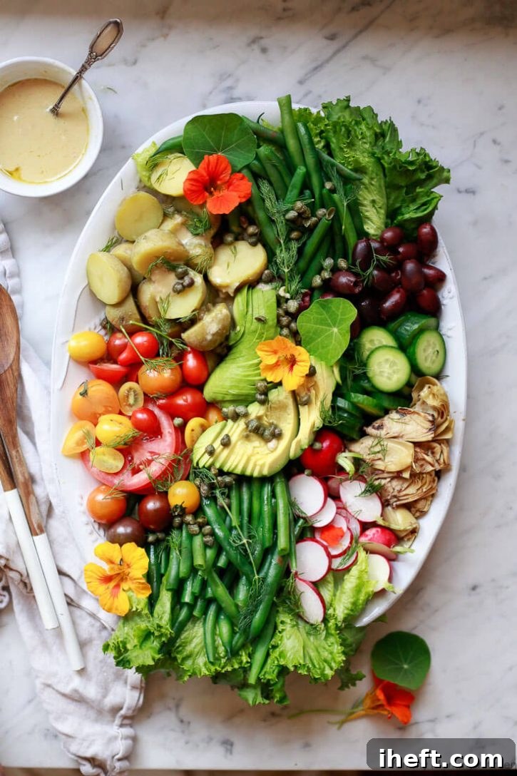 A beautiful Nicoise salad platter with green beans, potatoes, tomatoes, capers, olives, and avocado on a marble countertop with shallot vinaigrette on the side. 
