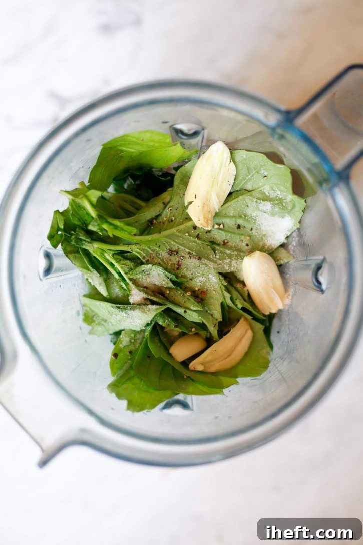 Fresh, vibrant spinach leaves, aromatic garlic cloves, and a selection of other key ingredients for a luscious avocado Green Goddess dressing are meticulously placed inside a powerful Vitamix blender, poised to be transformed into a smooth, creamy sauce.