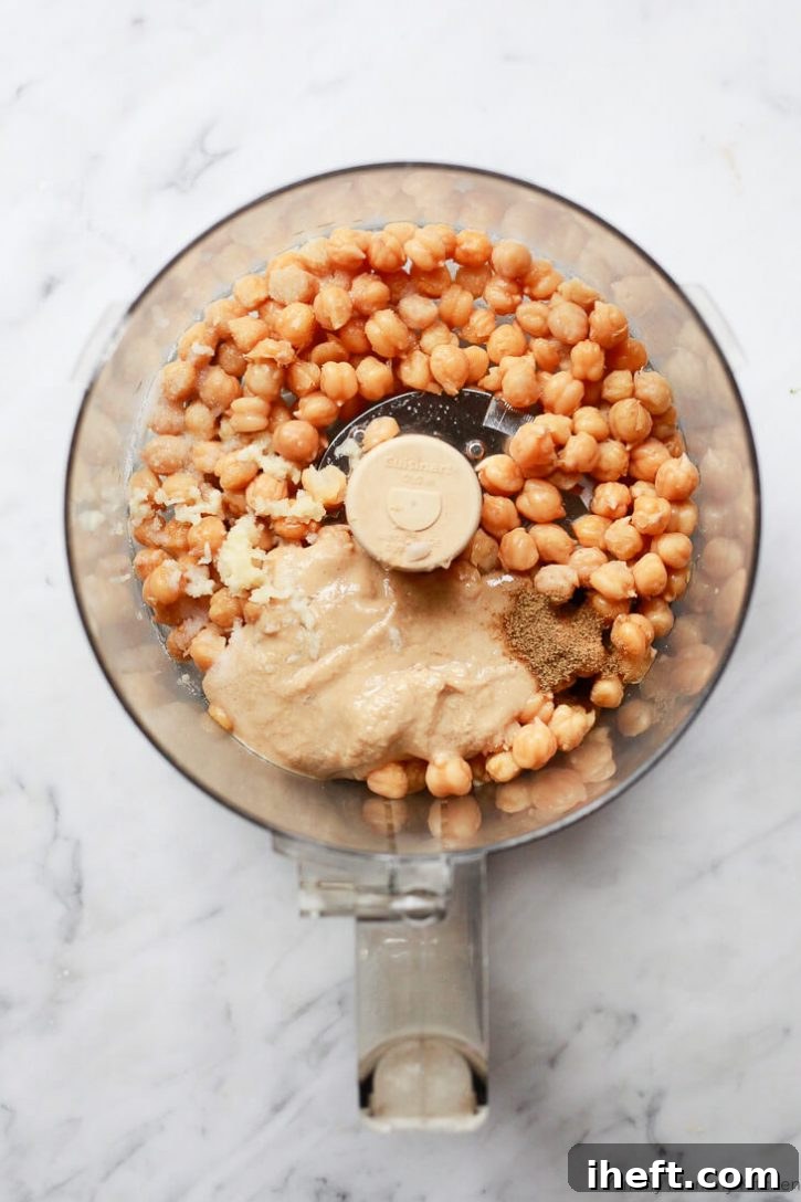A food processor filled with chickpeas, tahini, cumin, garlic, and lemon juice before blending into a homemade hummus recipe. 